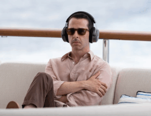 Kendall Roy sits crossed-arms and crossed-legs on a boat wearing dark glasses and headphones, clearly avoiding conversation or eye contact with anyone